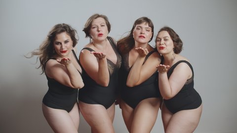 Stylish charming plus size models sending air kisses posing at camera. Four sexy overweight ladies in black lingerie isolated over grey background. Body positive and fashion concept
