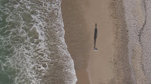 AERIAL DRONE TOP DOWN SHOT OF MAN WALKING ALONE IN THE SHORE OF THE BEACH WITH BIG SHADOW AND STRONG TIDE