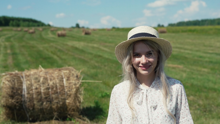 A charming portrait of a young country woman in white airy dress and hat. Woman stands in the field and smiliing in the camera.