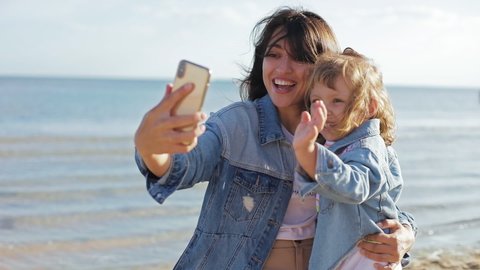 Mother and daughter talking online by video call on cellphone in summer sea. Portrait of smiling woman and girl having conversation by video chat outside. Happy family using smartphone