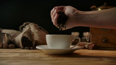Roasted arabica coffee beans pouring in coffee cup without processing. Woman's hand throws grains in slow motion. Concept for extra strong black coffee. Wood table, traditional coffee background วิดีโอสต็อก