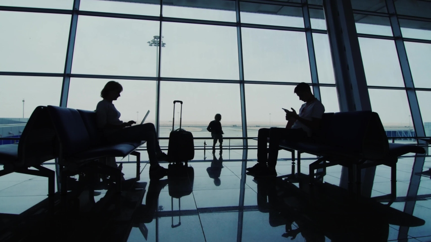 A woman, a man and a child in the airport waiting room. Silhouettes against the background of a large window | Shutterstock HD Video #1056644657