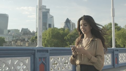 Pretty Young adult hispanic woman walking by over Tower Bridge using smart phone looking at city of London skyline
