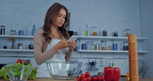 happy woman in apron using smartphone near ingredients on table