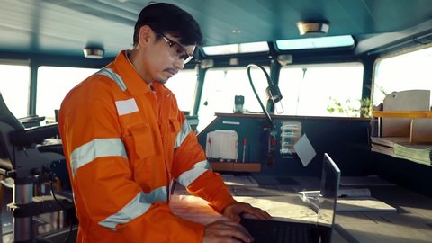 Filipino deck Officer on bridge of vessel or ship wearing coverall during navigaton watch at sea . He is using laptop, electronic paperwork at sea, concept of reporting