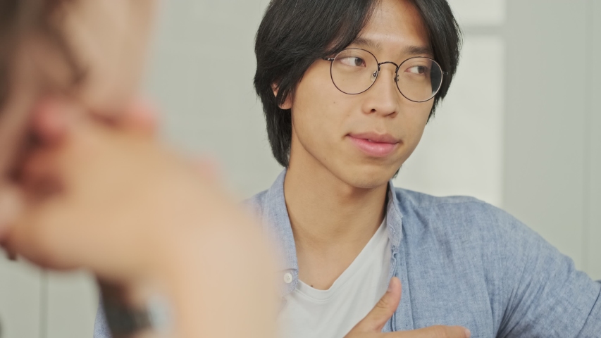 Calm young Asian man talking about something with his colleague while sitting by the table at the office. | Shutterstock HD Video #1056649094