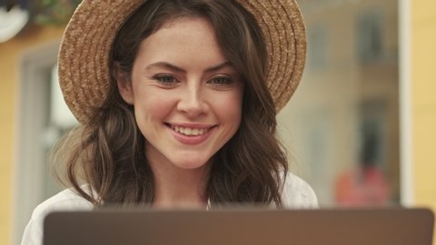 Smiling attractive woman wearing hat using laptop computer while sitting by the table in cozy cafe outdoors