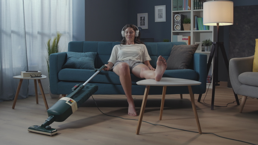 Lazy woman sitting on the couch and vacuuming the floor, she is bored and listening to music | Shutterstock HD Video #1056649676