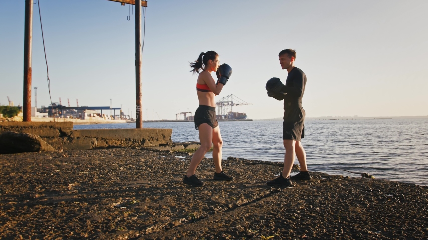 Young athletic lady in boxing gloves and sportswear is boxing a paw of her experienced trainer. They are training by seaside