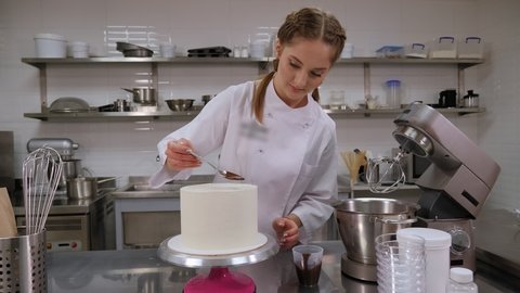 Close-up of a pastry chef pouring liquid chocolate from a spoon on a white cream sponge cake. Decorating a cake with cream and chocolate in a professional pastry kitchen.