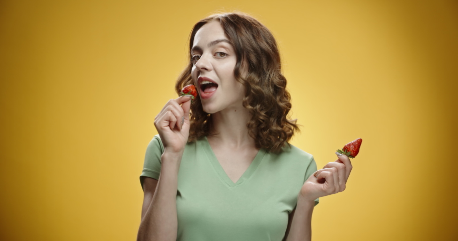 Positive caucasian lady eating strawberries and positively smiling, simbolizing healthy way of life, isolated on yellow background - emotions concept close up 4k