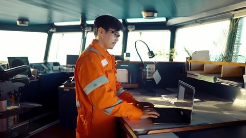 Filipino deck Officer on bridge of vessel or ship wearing coverall during navigaton watch at sea . He is using laptop, electronic paperwork at sea, concept of reporting