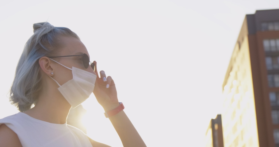 Beautiful Girl in Medical Mask Stands on City Street. Takes off Sunglasses and Looks Around. Close-up. Tourist after Pandemic. Sun Soft Lighting. Happy Girl. Ability to Travel. Summer. New Discoveries Royalty-Free Stock Footage #1056658937