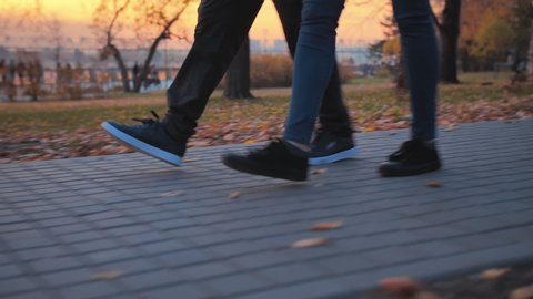 A young couple is walking in the autumn park against the backdrop of a beautiful sunset. Legs of two romantic people walking along the embankment against the background of people having a rest. Happy