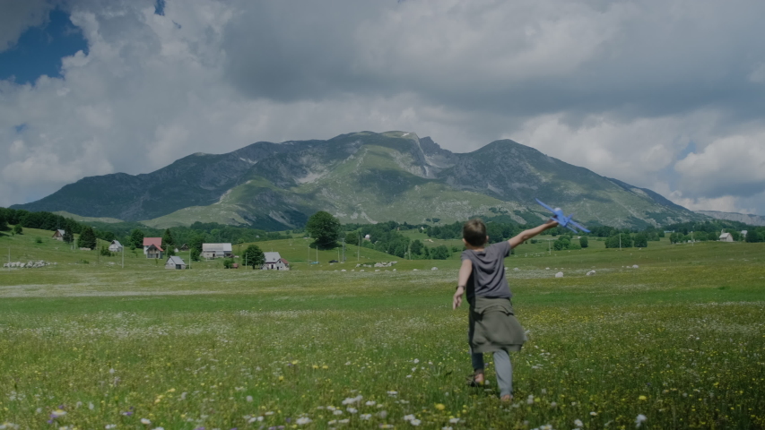  Cute boy kid with a toy blue plane runs through a meadow with flowers against a background of mountains in Montenegro. Happy childhood concept. 
