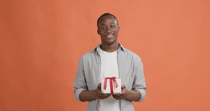Happy holidays. Cheerful african american guy reaching gift box to camera and winking, orange studio background