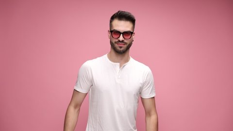 attractive macho casual man is flexing his muscles, kissing his biceps with pride, bowing his head and wearing sunglasses on pink background