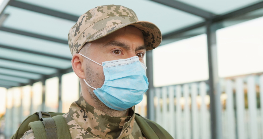 Portrait of serious Caucasian handsome young male soldier in cap and medical mask standing at street. Man militarian looking at camera at train station. Bus stop. Military uniform. Covid-19 pandemic. Royalty-Free Stock Footage #1056665408
