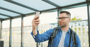 Caucasian handsome young man in glasses and with backpack standing at bus stop and talking on mobile phone via webcam. Male in eyeglasses having videochat on cellphone at train station. Videochatting.
