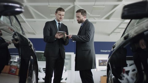 A male bearded client choosing car at dealership with the help of auto seller. Manager in auto dealership presents new cars using a tablet.