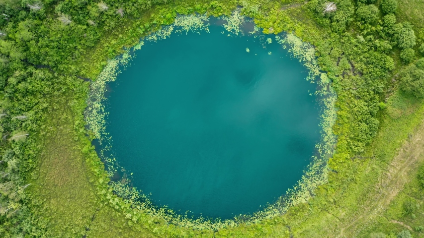 Aerial top down flight over amazing small lake of perfectly round shape. Cloudy sky reflected in clear turquoise water of pond surrounded by trees and plants. Untouched nature from above on summer day Royalty-Free Stock Footage #1056667406