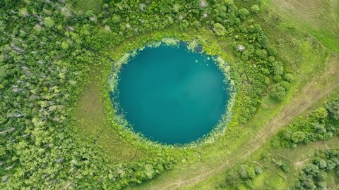 Aerial top down flight over amazing small lake of perfectly round shape. Cloudy sky reflected in clear turquoise water of pond surrounded by trees and plants. Untouched nature from above on summer day