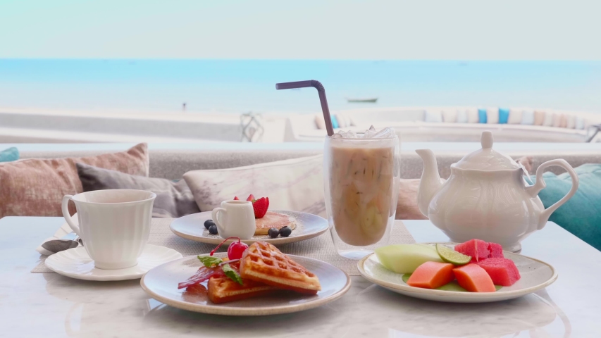 Outdoor Served Breakfast for Two on Table with Sea View. Glass of Iced Coffee, Cup of Tea, Teapot, Belgian Waffles, Pancake with Strawberry, Plate with Cutted Fruits. Hotel Sweet Lunch Concept. | Shutterstock HD Video #1056673400