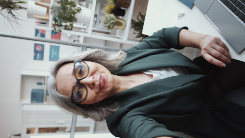 Vertical shot of attractive senior businesswoman looking at camera, waving and chatting online via video call on smartphone while sitting at office desk, a Business & Finance Video by AnnaStills