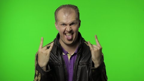 Happy rocker man in brown leather jacket showing rock and roll sign, devil horns gesture and looking with crazy expression. Portrait of guy biker posing on chroma key background. People emotions
