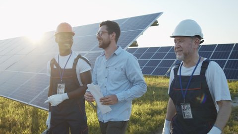 Cheerful multi-racial workers with engineer walking on solar farm. Successful business. Ecological construction. Solar panels field. Teamwork.
