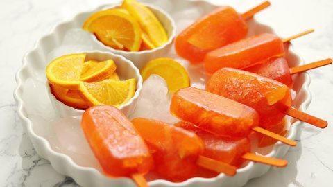 Homemade, juicy, orange popsicles. Placed on a white plate with ice cubes. Flat lay on white marble with fresh orange slices. Top view.