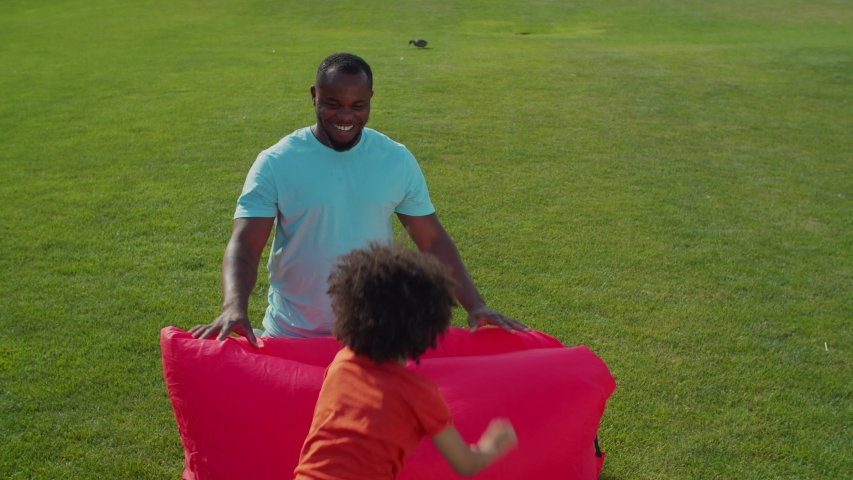 Positive smiling black father with carefree adorable little son having fun outdoors, fooling around on air lounger on park lawn, playing leisure games and bonding during weekend on summer day.