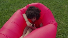 Portrait of smiling happy cute african american preschooler boy with curly hair busy playing video games online on cellphone, resting on inflatable lounger while enjoying leisure in public park.