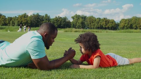 Joyful handsome african american father and playful adorable little mixed race son with curly hair arm wrestling lying on green grass in public park, enjoying playtime and leisure in summer nature.