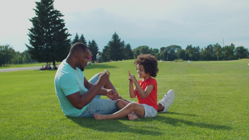 Positive black father with adorable preschooler mixed race son with curly hair sitting on green grass, having fun playing rock paper scissors game while enjoying leisure together in summer nature.