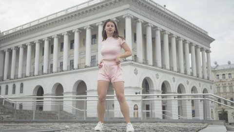 Wide shot of beautiful twerk dancer performing outdoors on city square. Portrait of cheerful brunette Caucasian woman dancing modern dance at the background of ancient European building.