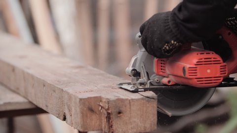 Carpenter in work clothes and gloves neatly cuts piece of board with circular electric saw and strokes wood with hand, close-up view in slow motion. Flying sawdust of working circular. 