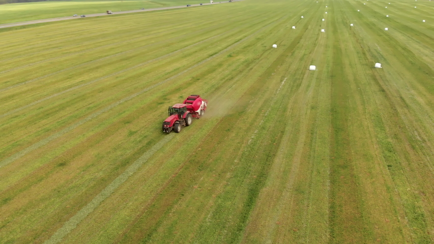 A tractor with a press collector collects and presses dry straw into a white polymer film for feeding livestock in winter. Packed bales of hay on a green field, top view