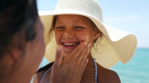 Close up of young mother is applying protective sunscreen or sunblock lotion on her little happy smiling daughter's face to take care of skin on a seaside beach during family holidays vacation.
