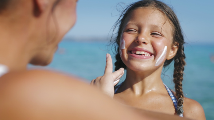 Close up of young mother is applying protective sunscreen or sunblock lotion on her little happy smiling daughter's face to take care of skin on a seaside beach during family holidays vacation. | Shutterstock HD Video #1056688361