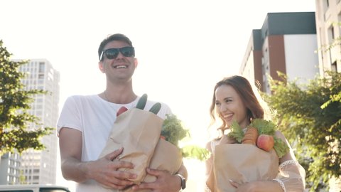 Happy Carefree Couple Laughs Walking Along Street to Home from Market, Carrying Bags with Fresh Healthy Food. Fun Grocery Shopping Together, Casual Life of Young Excited Family Enjoying Joke Outdoors