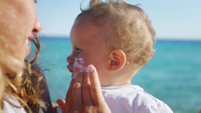 Close up of young mother is applying a protective sunscreen or sunblock lotion on her little toddler son face to take care of his delicate skin on a seaside beach during family holidays vacation. | Shutterstock HD Video #1056689267