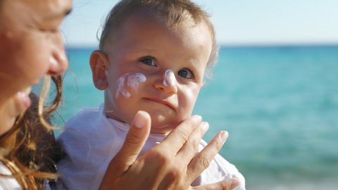 Close up of young mother is applying a protective sunscreen or sunblock lotion on her little toddler son face to take care of his delicate skin on a seaside beach during family holidays vacation.