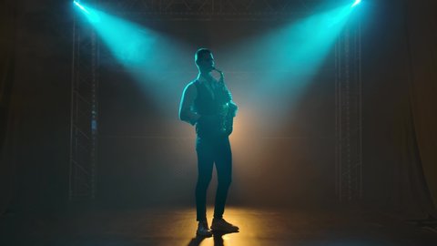 Silhouette a young stylish guy plays the golden shiny saxophone in the turquoise spotlights on stage. Dark studio with smoke and neon lighting. Neon lighting effects. Side view. Slow motion.