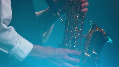 A young retro stylish guy plays on the golden shiny saxophone in the turquoise spotlights on stage. Dark studio with smoke and neon lighting. Hands and saxophone close up. Side view.