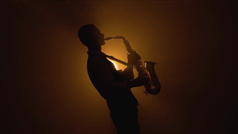 Silhouette a young stylish guy plays the golden shiny saxophone in the yellow spotlights on stage. Dark studio with smoke and neon lighting.