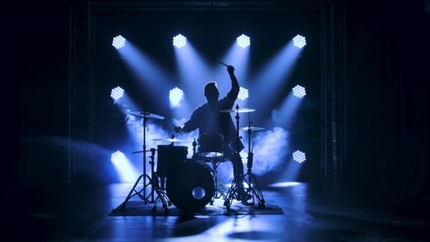 Silhouette drummer playing on drum kit on stage in a dark studio with smoke and neon lighting. Dynamic neon lighting effects. Performance vocal and musical band. Slow motion.