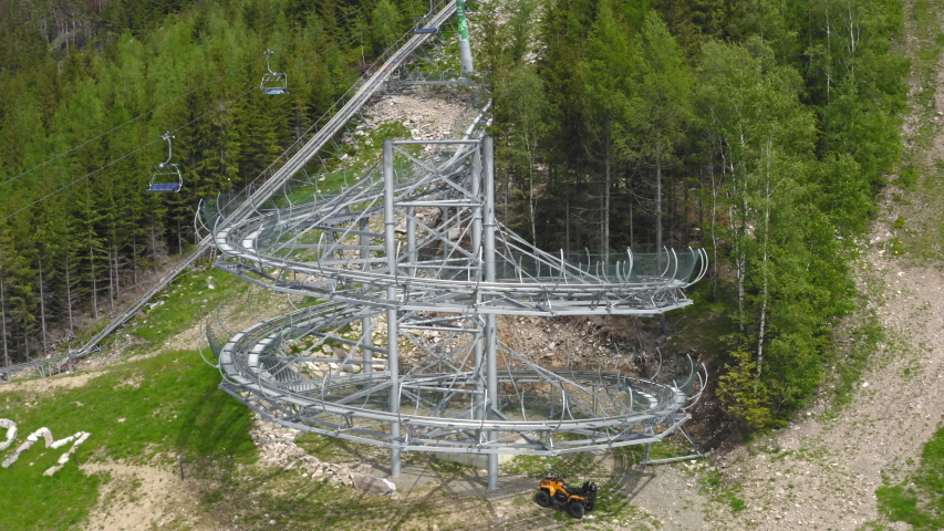 Rotating aerial 4k shot of a cart with people spiralling down a big loop of an extreme outdoor roller coaster in the mountains of Dolní Morava, Czech Republic with trees around. Mamutí horská dráha.