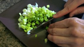 A woman is quickly chopping fresh spring onions (scallions) on a plate using a sharp knife. A close up footage suitable for demonstration of home cooking, healthy eating, vegan or vegetarian diets.