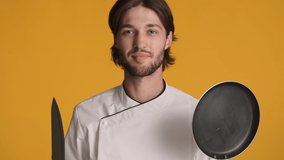 Professional excited chef in uniform holding knife and frying pan surprisingly looking in camera over colorful background
